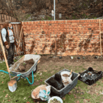 Sadhana Kraus bei der Arbeit an einer Backsteinmauer, davor Schubkarre, Werkzeug und Kisten mit Erde