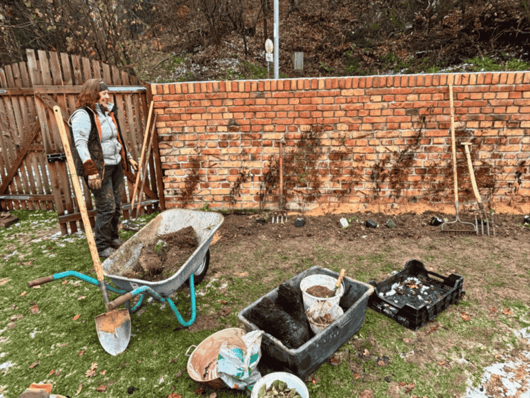 Sadhana Kraus bei der Arbeit an einer Backsteinmauer, davor Schubkarre, Werkzeug und Kisten mit Erde