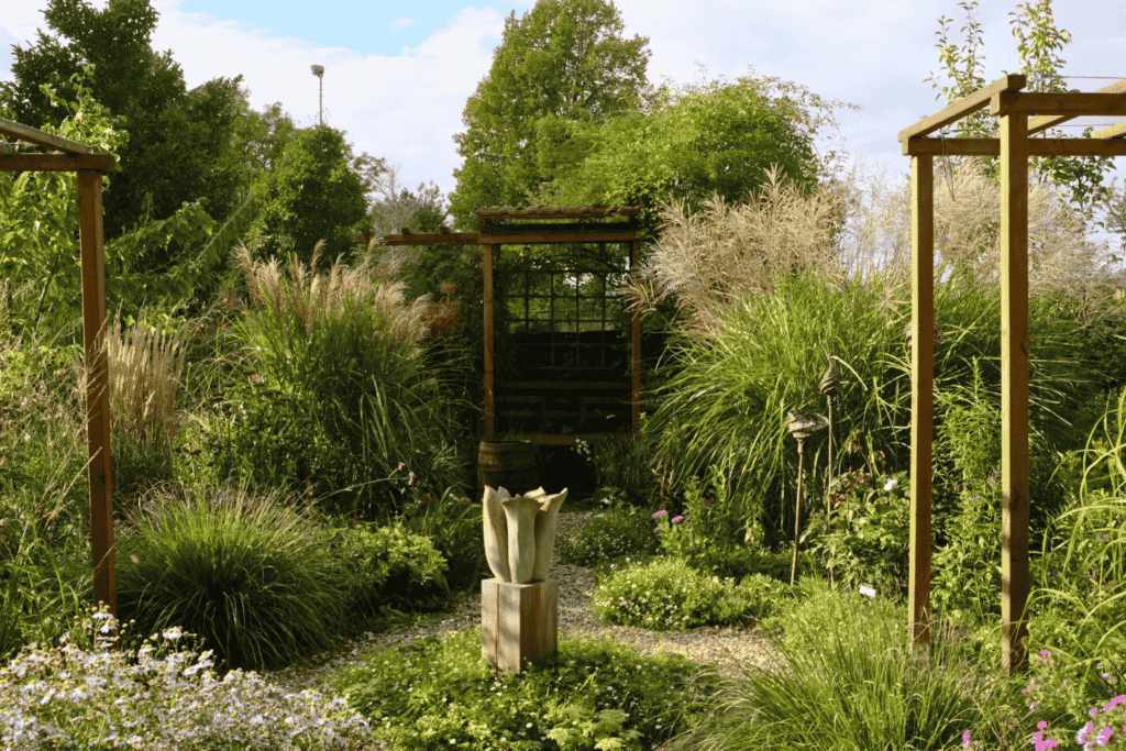 Staudengarten mit Kieswegen, flankiert von üppigen Gräsern und Stauden, der auf eine zentrale Skulptur und eine begrünte Pergola als Blickfang zuläuft