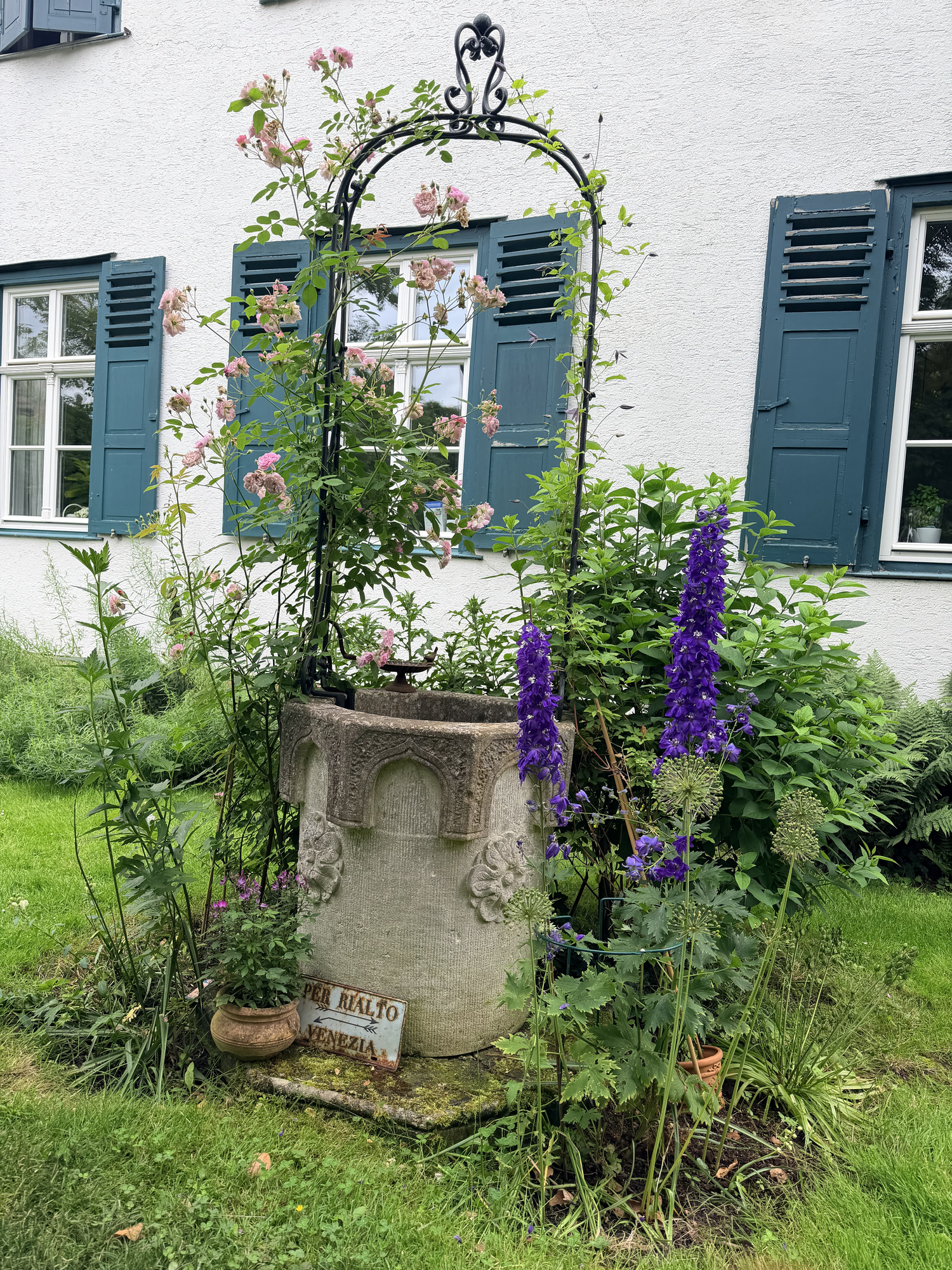 Venezianischer Brunnenstein mit Rosenbogen und blauem Rittersporn vor dem Haus
