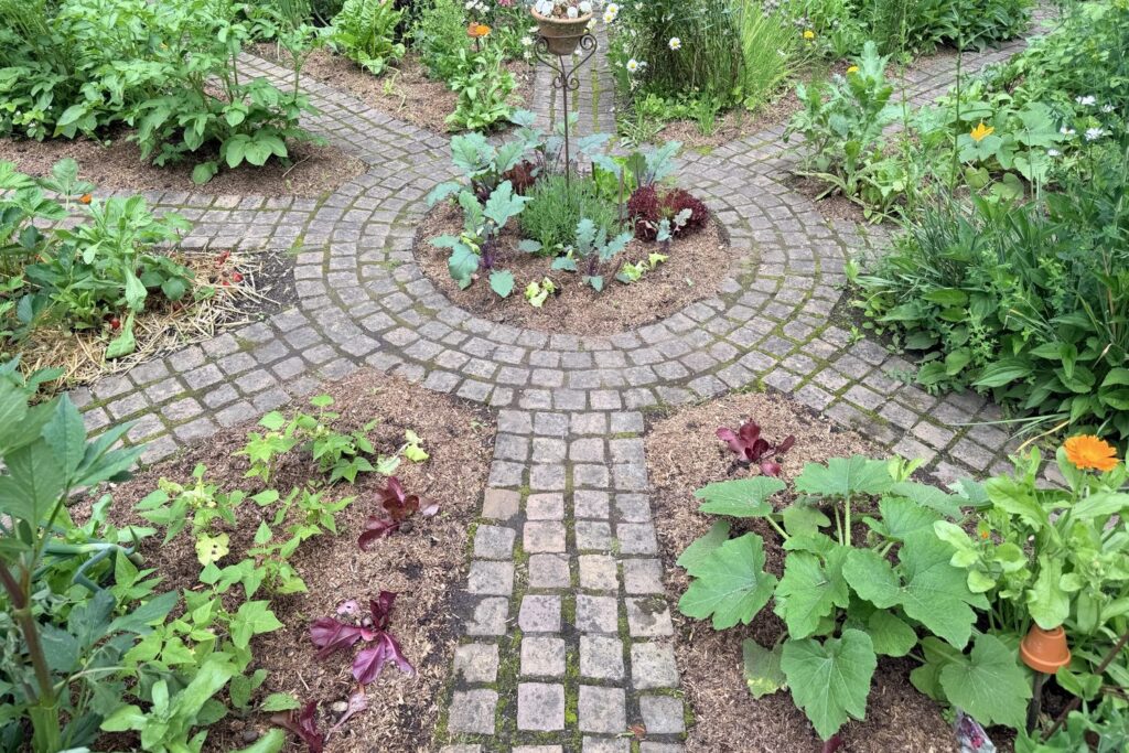 Wegekreuz aus Pflastersteinen im Bauerngarten mit Salat, Zucchini und Ringelblumen (Calendula officinalis)